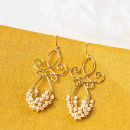Polished gold-tone/pearl white dangle earring with an elegant looped pattern, fine intricate detailing, and delicate small pearl bead embellishments, secured with a French hook closure.