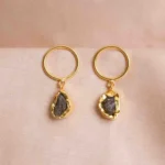 Handcrafted metal dangle earrings featuring a circular disc and a dangling raw teardrop gemstone in brownish-grey with golden and moss green undertones, secured with a push-back closure.