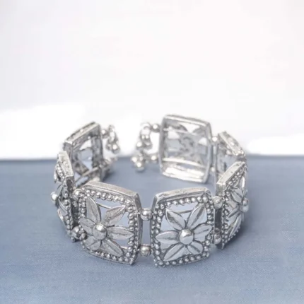 Handcrafted oxidized silver-tone metal cuff bracelet with evenly spaced floral square engravings, circular beadwork, and delicate ghungroo dangles.