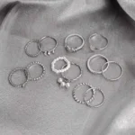 A large collection of twelve silver-toned stacking rings, including bands with pearl drops, continuous faux pearls, twisted rope texture, single crystal accents, and plain polished metal, scattered on a dark surface.