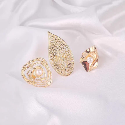 A trio of large, gold-toned statement rings. The set includes a wide etched leaf-shaped ring, a swirling abstract ring that cradles a pearl, and an asymmetrical eye-shaped ring also set with a pearl.