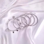 Set of six silver bangles with polished, twisted, and beaded designs, including a heart charm detail, crafted in lightweight slip-on style for versatile wear.