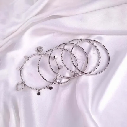 Set of six silver bangles with polished, twisted, and beaded designs, including a heart charm detail, crafted in lightweight slip-on style for versatile wear.
