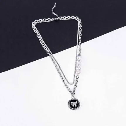 A silver-toned layered necklace is displayed on a dark gray laptop keyboard. The necklace features two main strands: one is a chunky curb chain with a circular pendant depicting a silver teddy bear, and the other is a finer chain adorned with five evenly spaced, lustrous white pearls. Both chains connect at the clasp end.